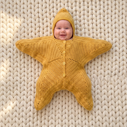 Smiling baby wearing the Nova Star Suit in butter yellow, lying on a cream chunky-knit blanket — hooded crochet star suit with buttoned front