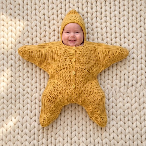 Smiling baby wearing the Nova Star Suit in butter yellow, lying on a cream chunky-knit blanket — hooded crochet star suit with buttoned front