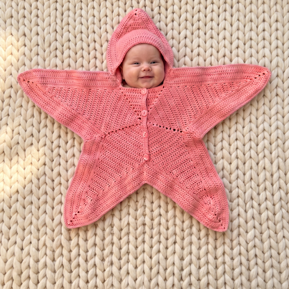 Baby wearing the Nova Star Suit crochet pattern in pink on a cream knit blanket, hood up, all five star points showing